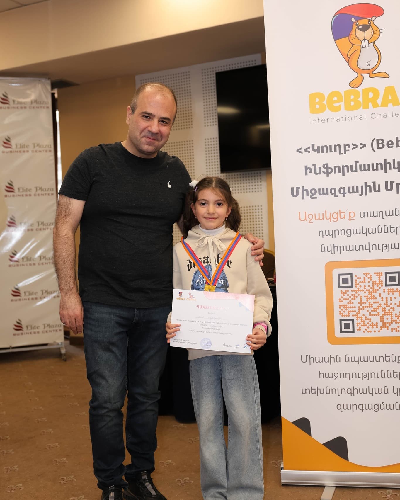Девочка с медальной лентой держит свой сертификат Bebras, стоя рядом с Ашотом Тонояном.