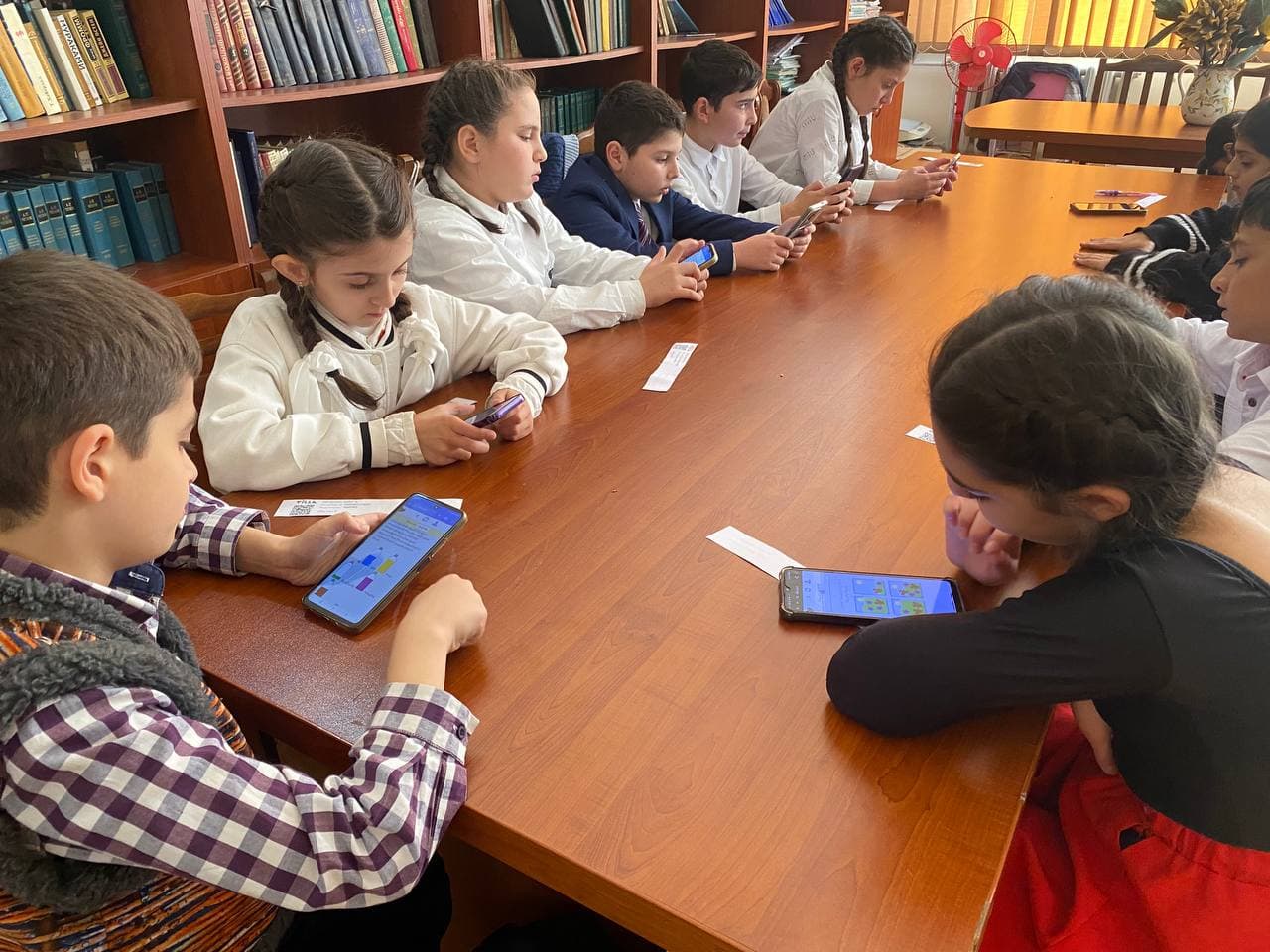 Ten children along a long library table under floor-to-ceiling shelves, every one of them working.
