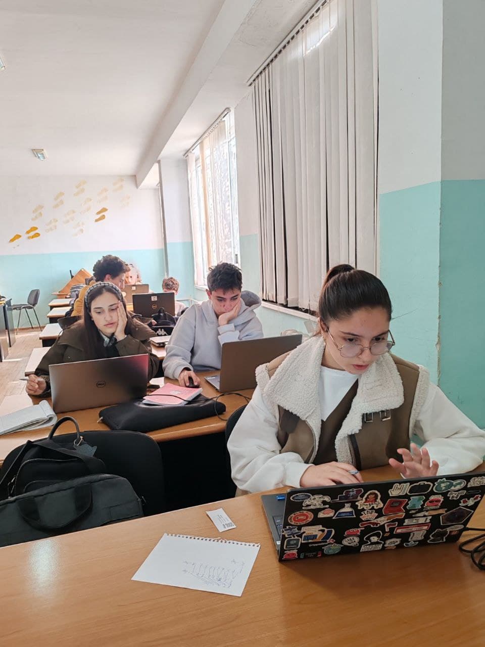 A student pauses mid-task, one hand raised, working through a Bebras problem on a laptop.