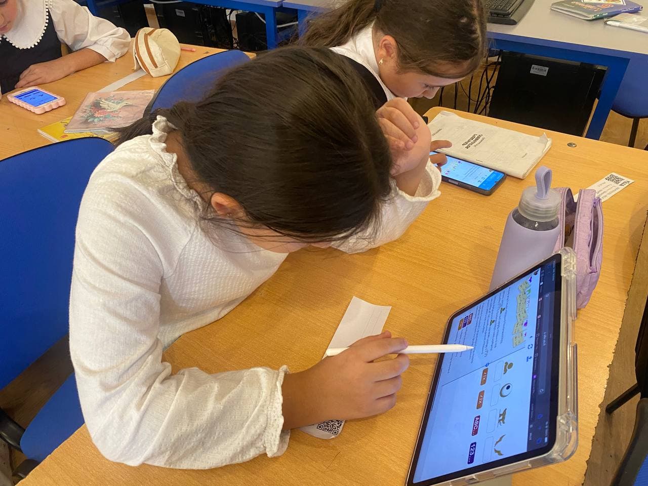 Two students bent over a tablet, one with a stylus hovering over a Bebras task.
