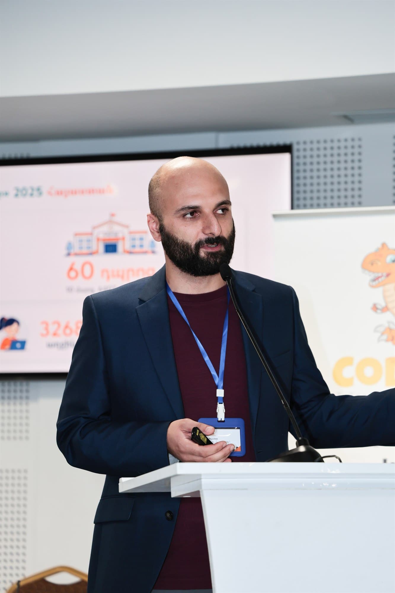David Khachatryan speaking at a lectern at the Bebras award ceremony, the year’s results on the screen behind him.