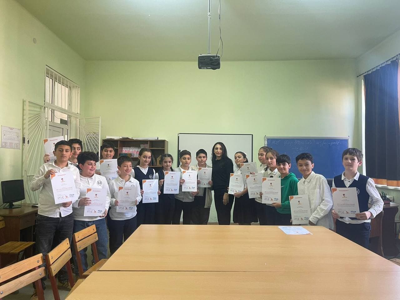 Класс детей в школьной форме с напечатанными сертификатами Bebras, их учительница стоит среди них у доски.